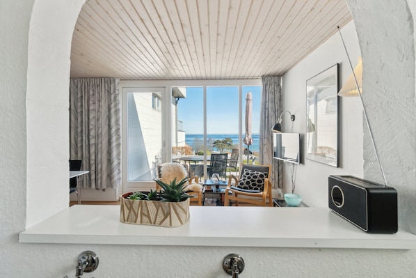 A modern interior with a white ceiling, a view of the sea, a white countertop, a black radio, and a plant in a geometric pot.