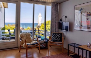 Habitación con un amplio ventanal corredero que da a un balcón con vistas al mar. Hay dos sillones, una mesa y un televisor montado en la pared.