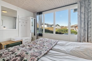 A bedroom with a large bed, a wardrobe, a mirror, and a window with a view of houses.