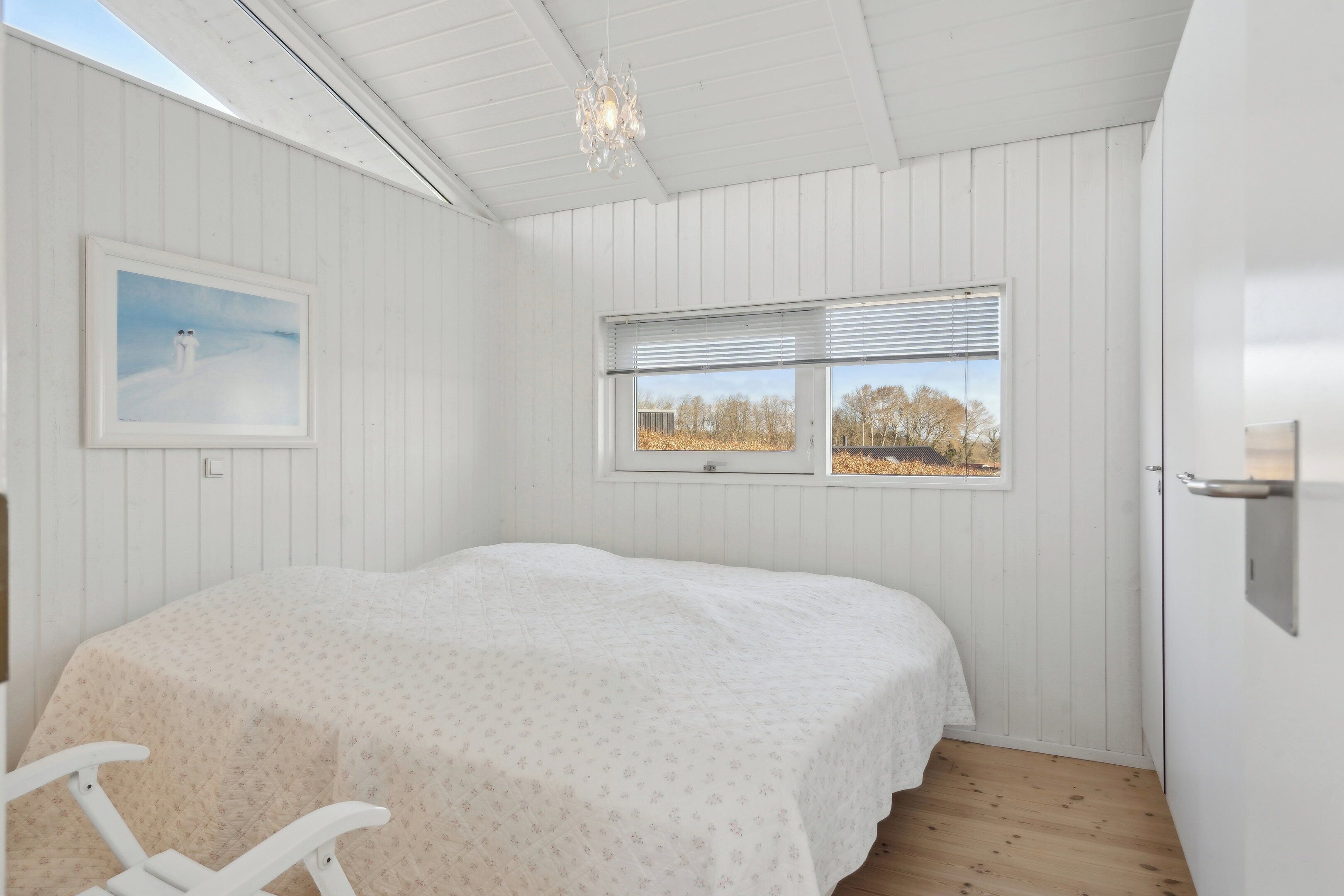 A bedroom with a bed, a window with blinds, a picture on the wall, and a chandelier.