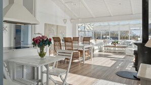 A bright, open-plan living area with a dining table, wicker chairs, a white sofa, and a large window providing a view of the outdoors. - Lovely summer house with sea view (Sjølund)