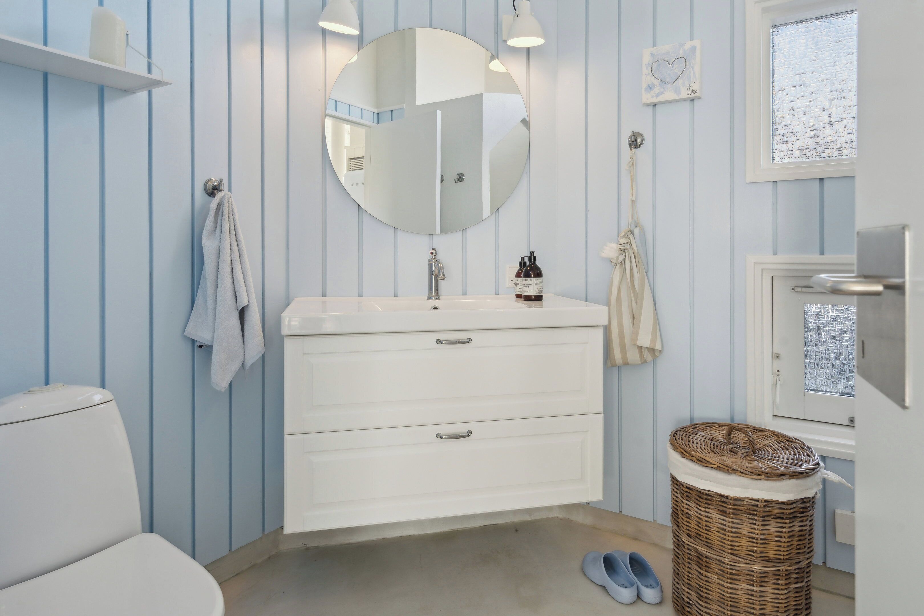 A bathroom with a white sink, mirror, and towel rack; a wicker basket; and a window with a view.