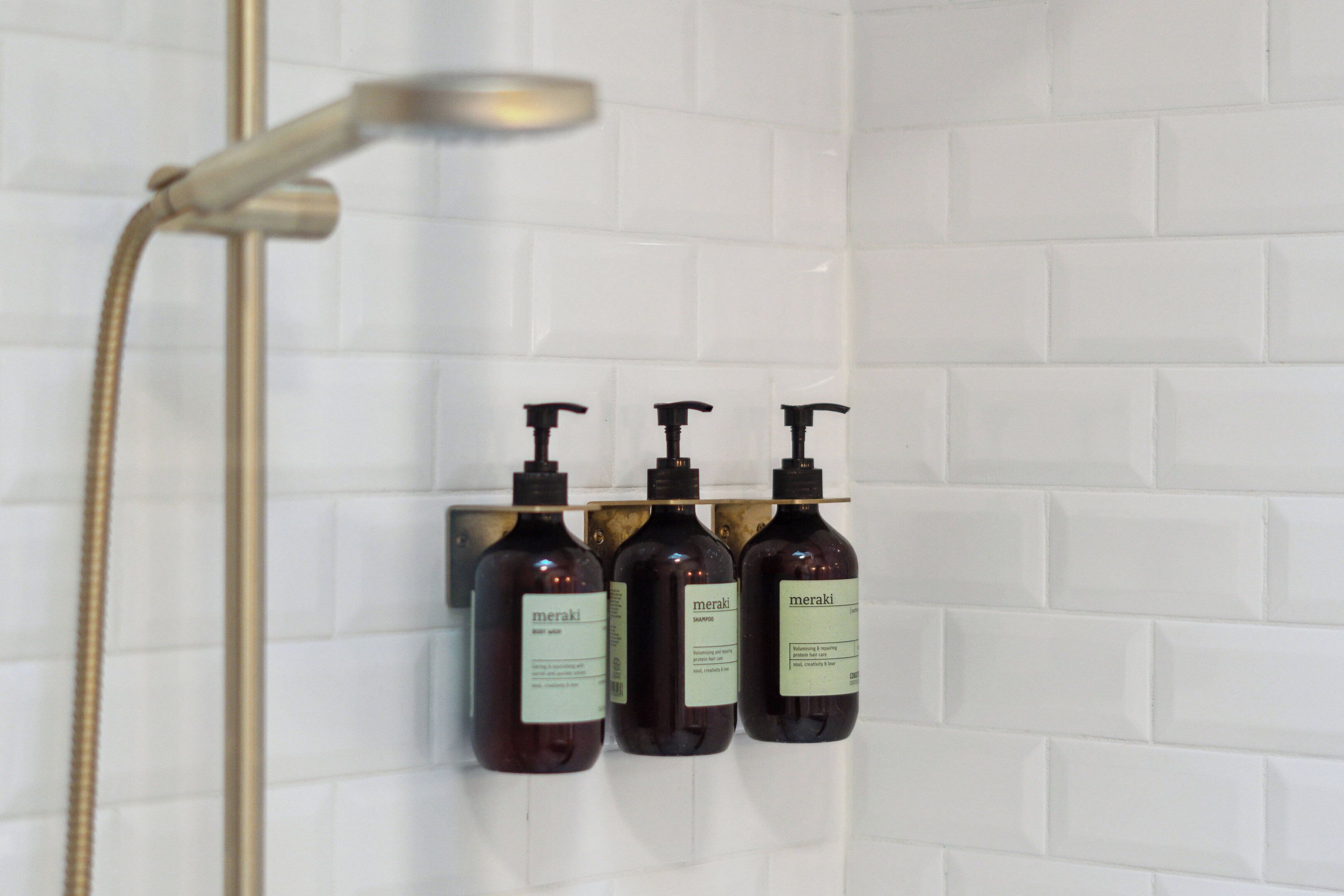 Ducha con cabezal de ducha manual y tres botellas de champú Meraki sobre una pared con azulejos.