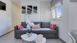 A modern living room with a grey sofa, white coffee table, and a small side table. There are three framed pictures on the wall, a ceiling fan, and a window with blinds. - Bellavista Studio (Albufeira)