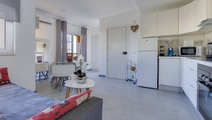 A modern, well-lit room with a bed, a gray sofa, a dining table with chairs, a kitchen area with white cabinets, a microwave, and a refrigerator. - Bellavista Studio (Albufeira)