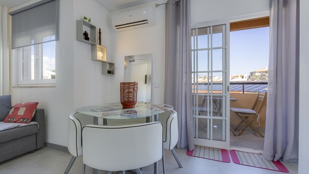 A modern living room with a glass table, white chairs, a grey sofa, a red cushion, and a sliding glass door leading to a balcony with solar panels. - Bellavista Studio (Albufeira)
