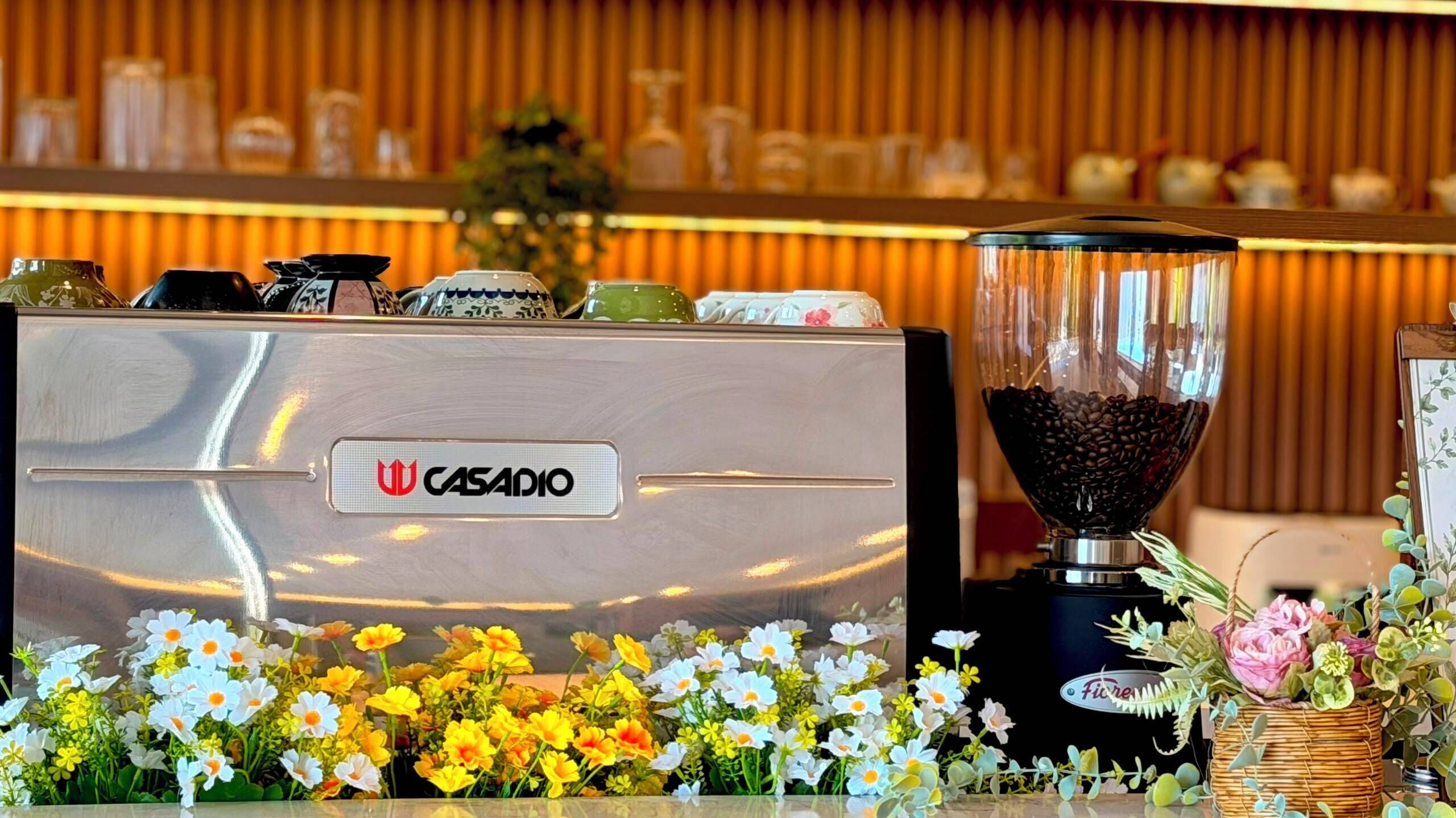A coffee machine branded CASADIO with a coffee grinder beside it, surrounded by a variety of flowers.