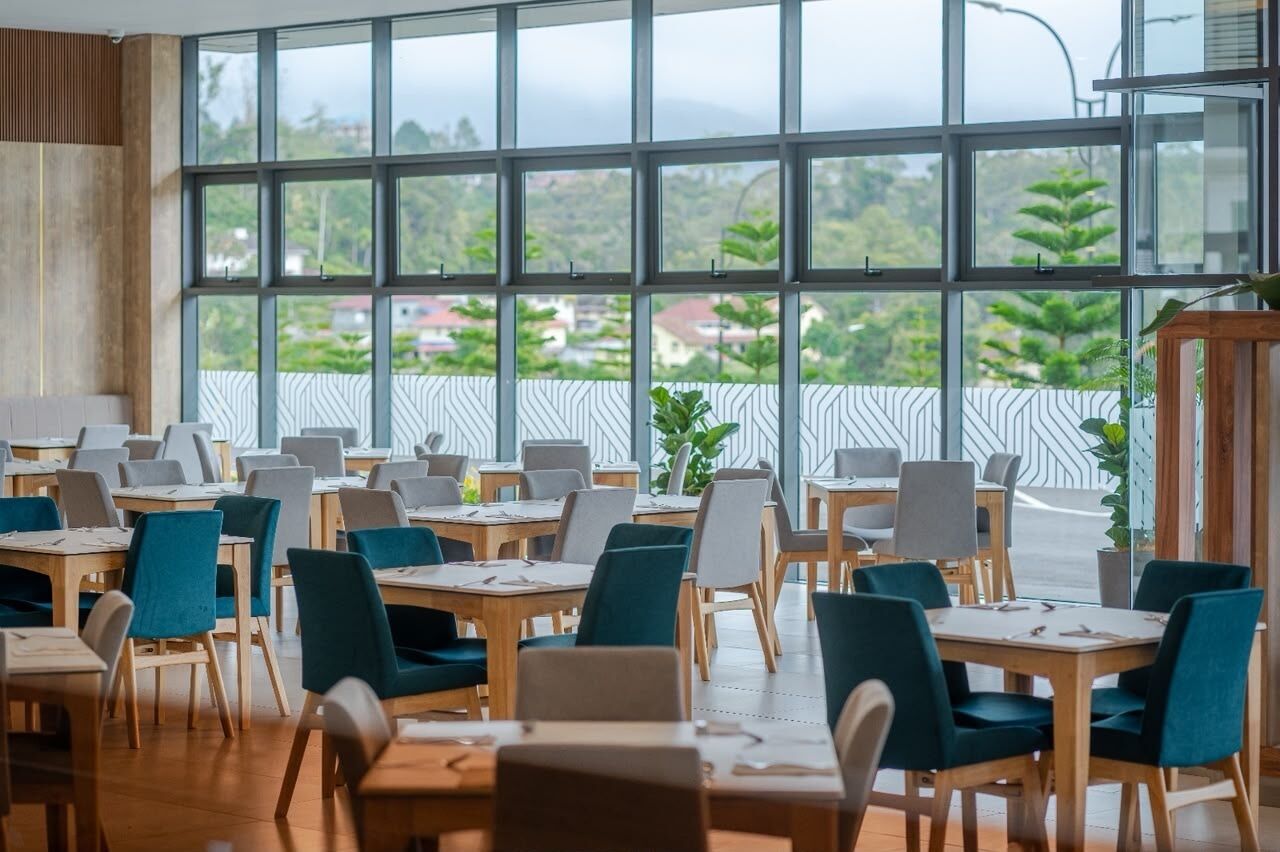 A dining area with multiple tables and chairs, large windows with a view of greenery and buildings, and a patterned railing along the window.