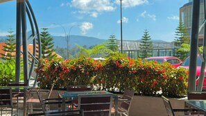 An outdoor seating area with tables and chairs, surrounded by greenery and a view of a bridge and mountains.