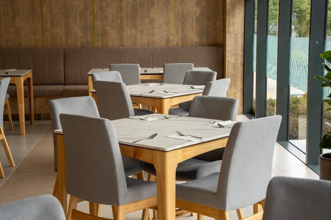 A dining area with a long wooden table and grey upholstered chairs, a wooden bench, and a view through large windows.