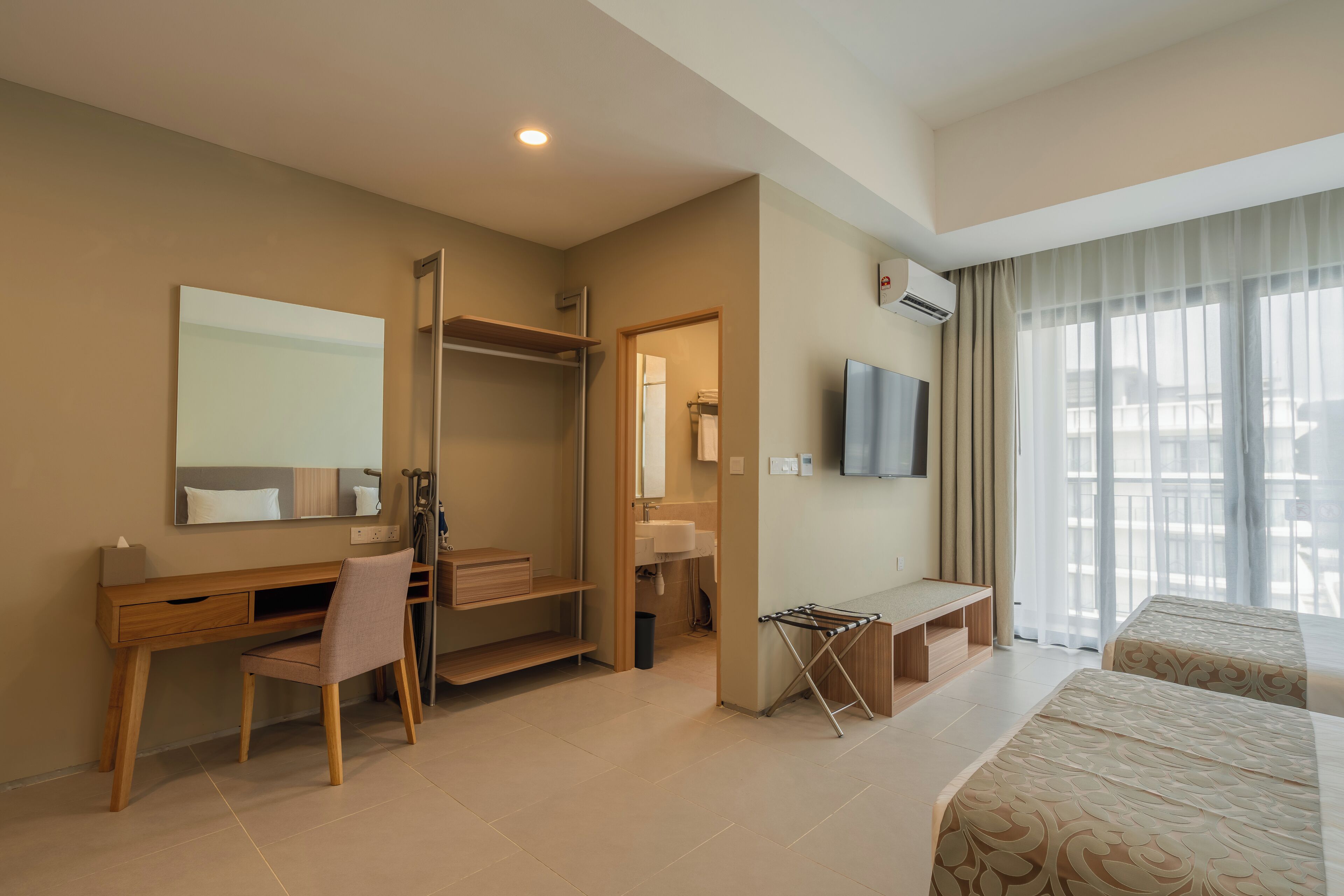 A hotel room with a wooden desk, a chair, a TV, a bed, a window with curtains, and a bathroom with a mirror and sink.
