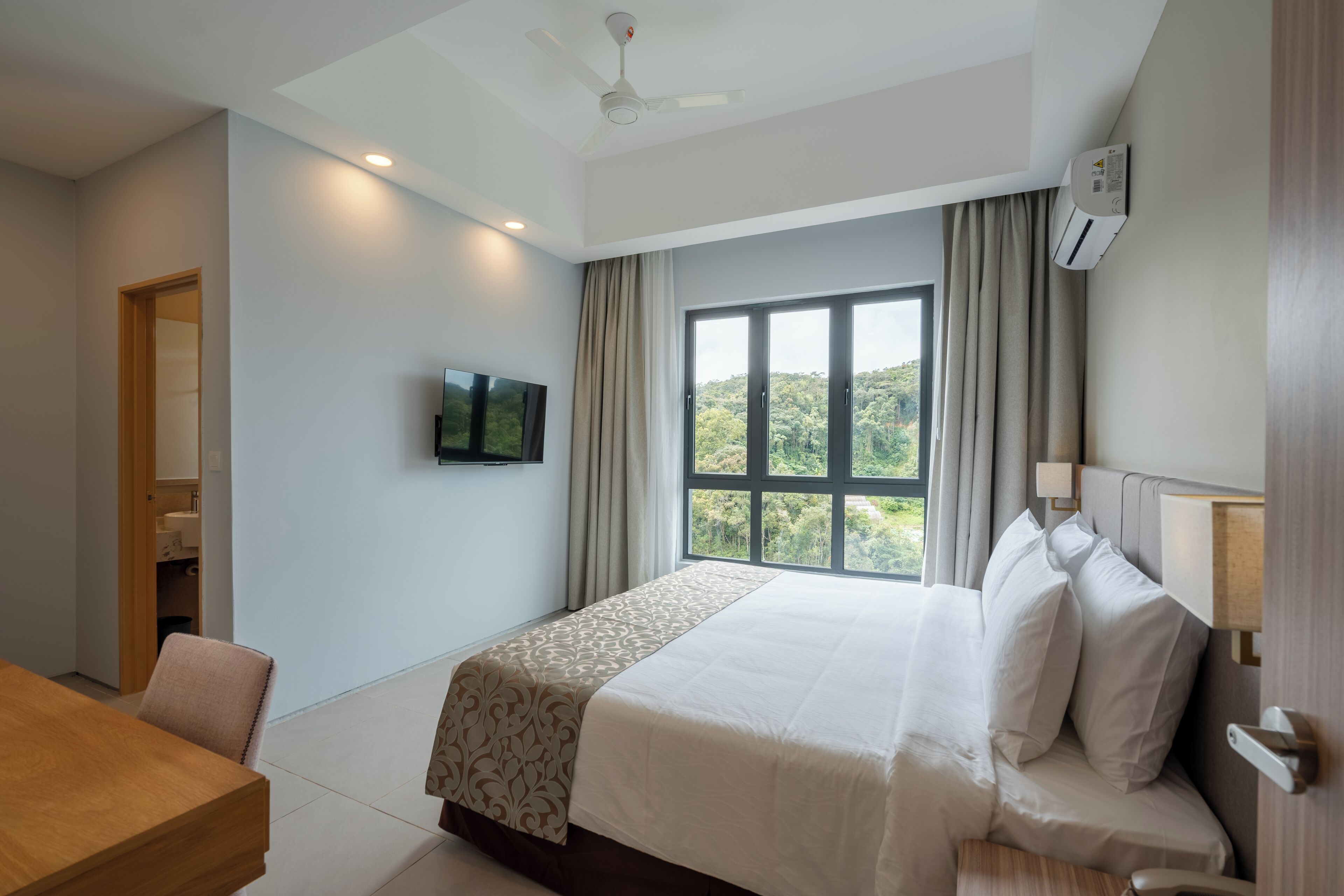 A hotel room with a bed, a TV mounted on the wall, a window with curtains, a ceiling fan, and an air conditioning unit.