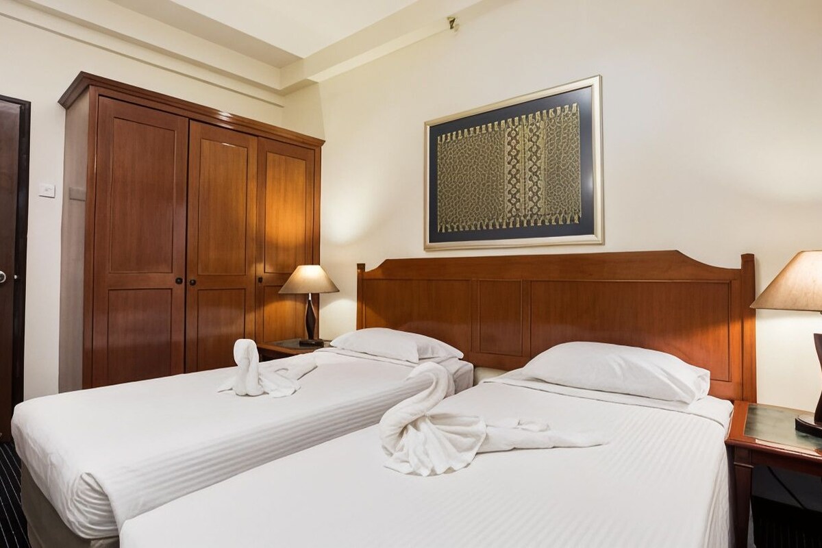 A hotel room with two beds, wooden headboards, a large wooden wardrobe, a framed decorative piece on the wall, and a nightstand with a lamp.