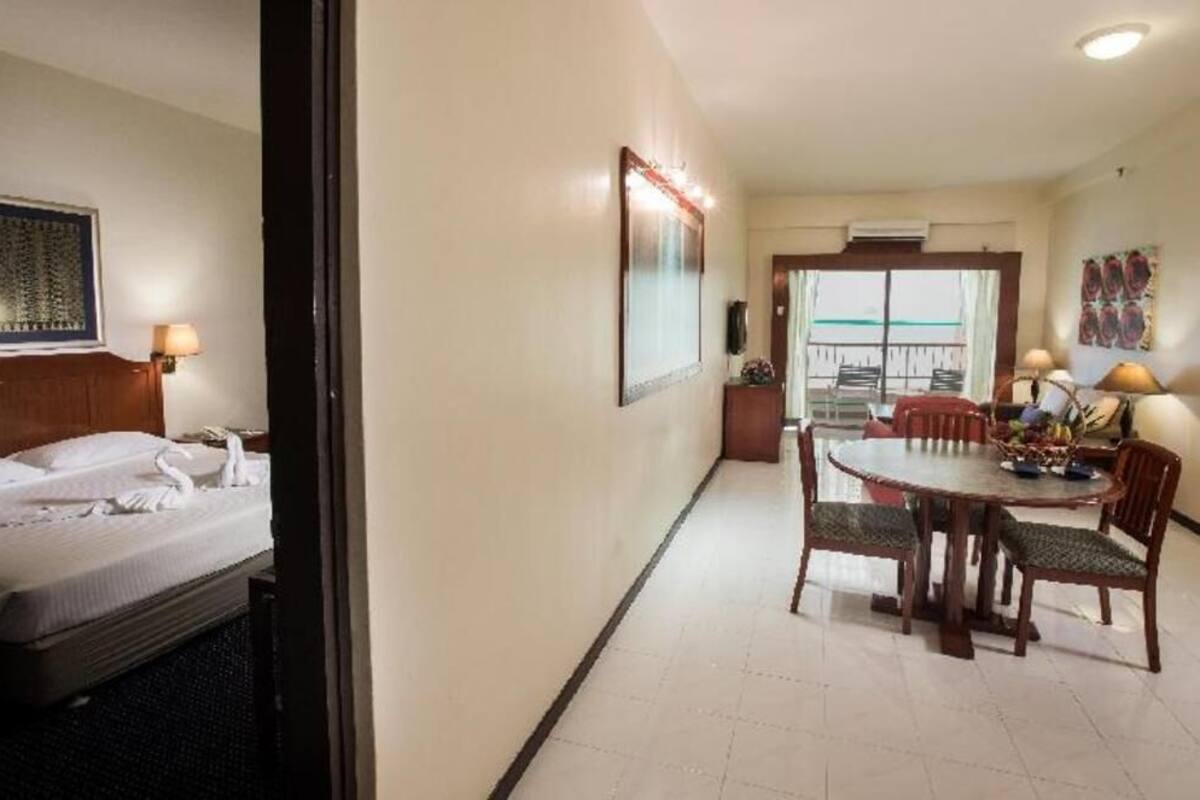 A hotel room with a bed, a dining table with chairs, a balcony with chairs, and a wall-mounted TV.