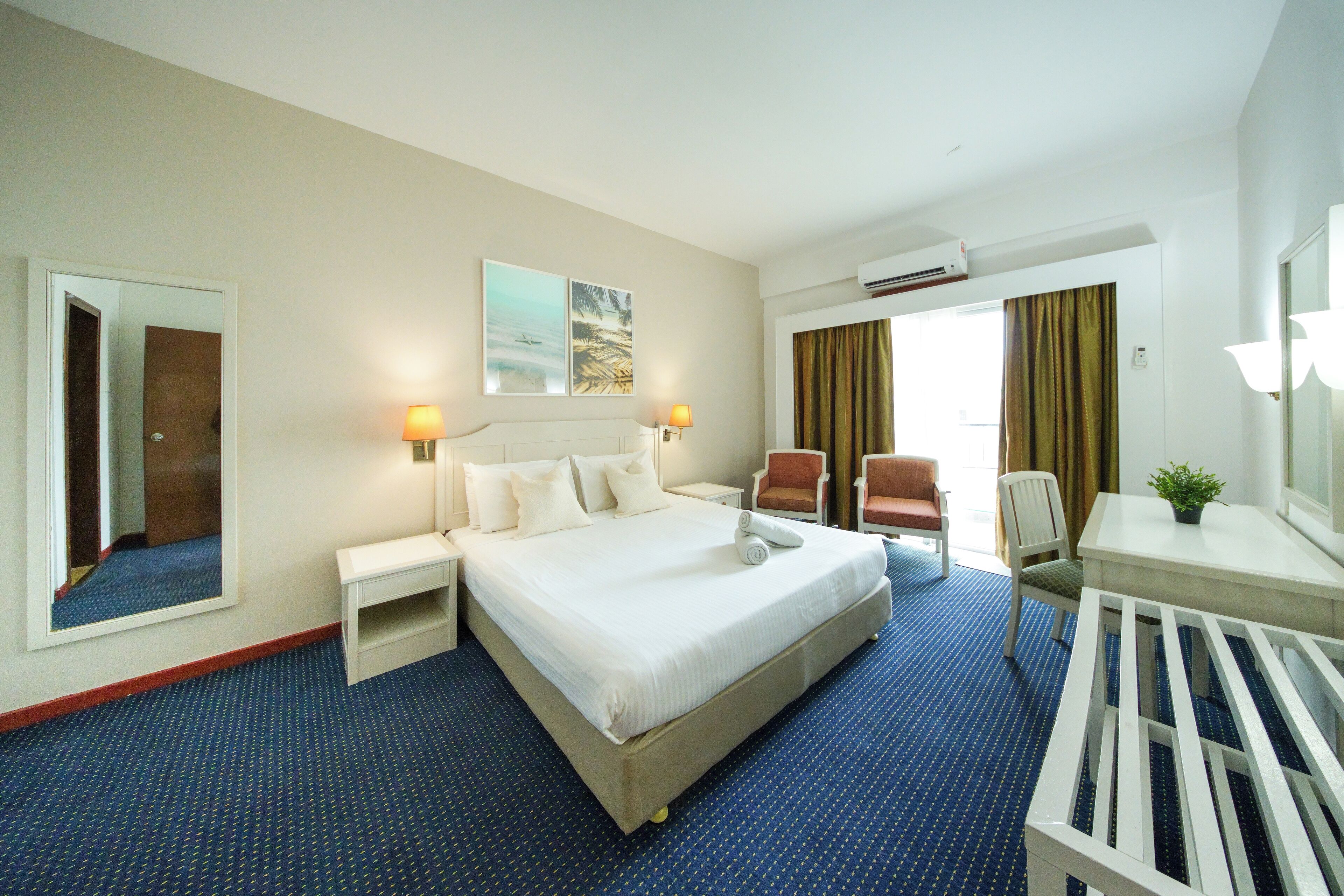 A hotel room with a large bed, bedside tables, a desk with a chair, a small table with a plant, and a mirror.