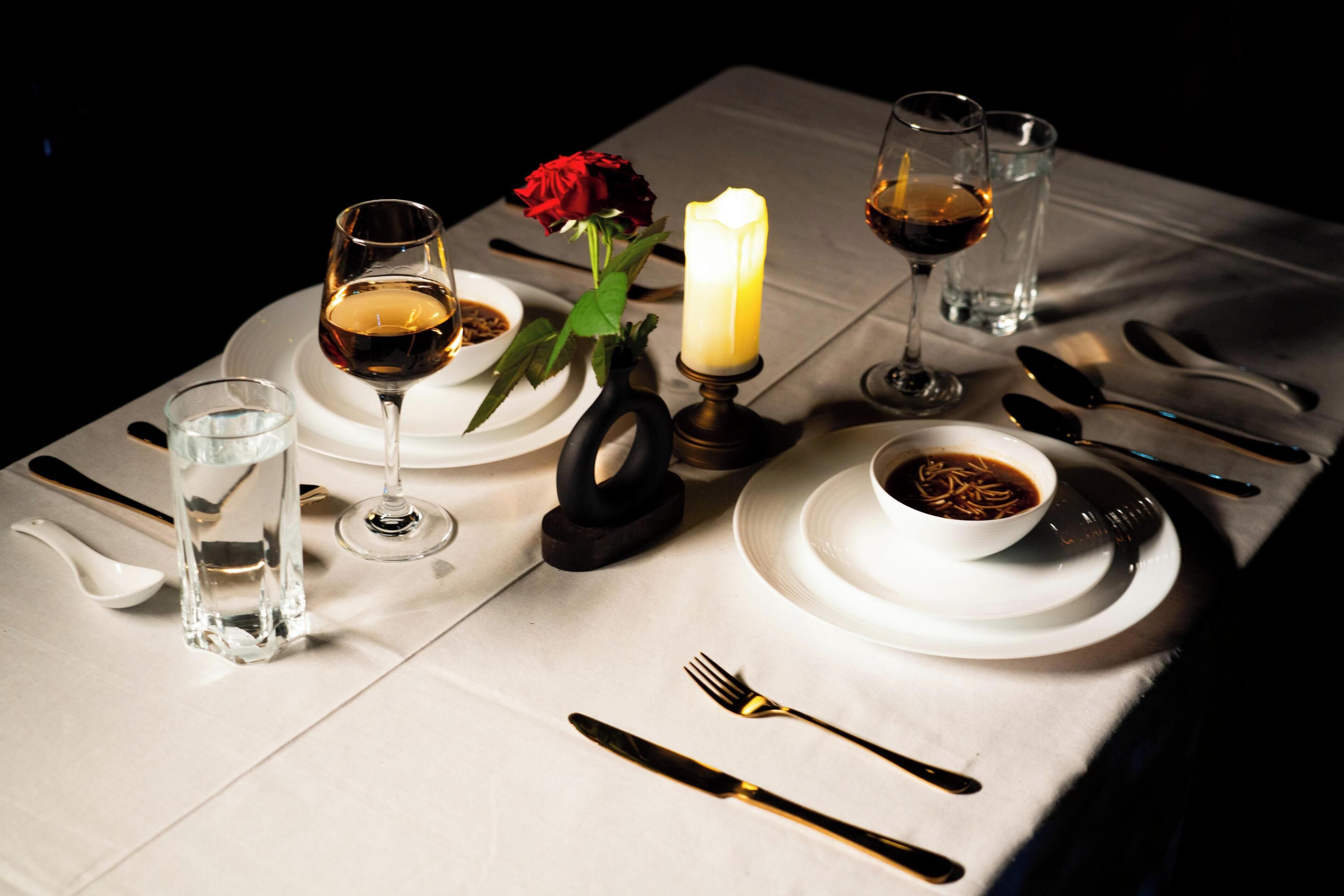 A table set for a meal with wine glasses, a candle, a rose, and a bowl of soup.
