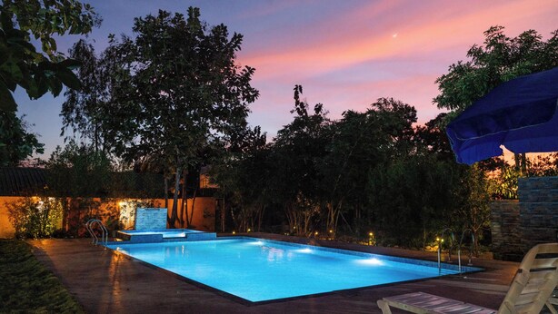 An outdoor swimming pool at dusk with surrounding trees and a stone wall. - Wagharanya Resort (Bhadravathi)