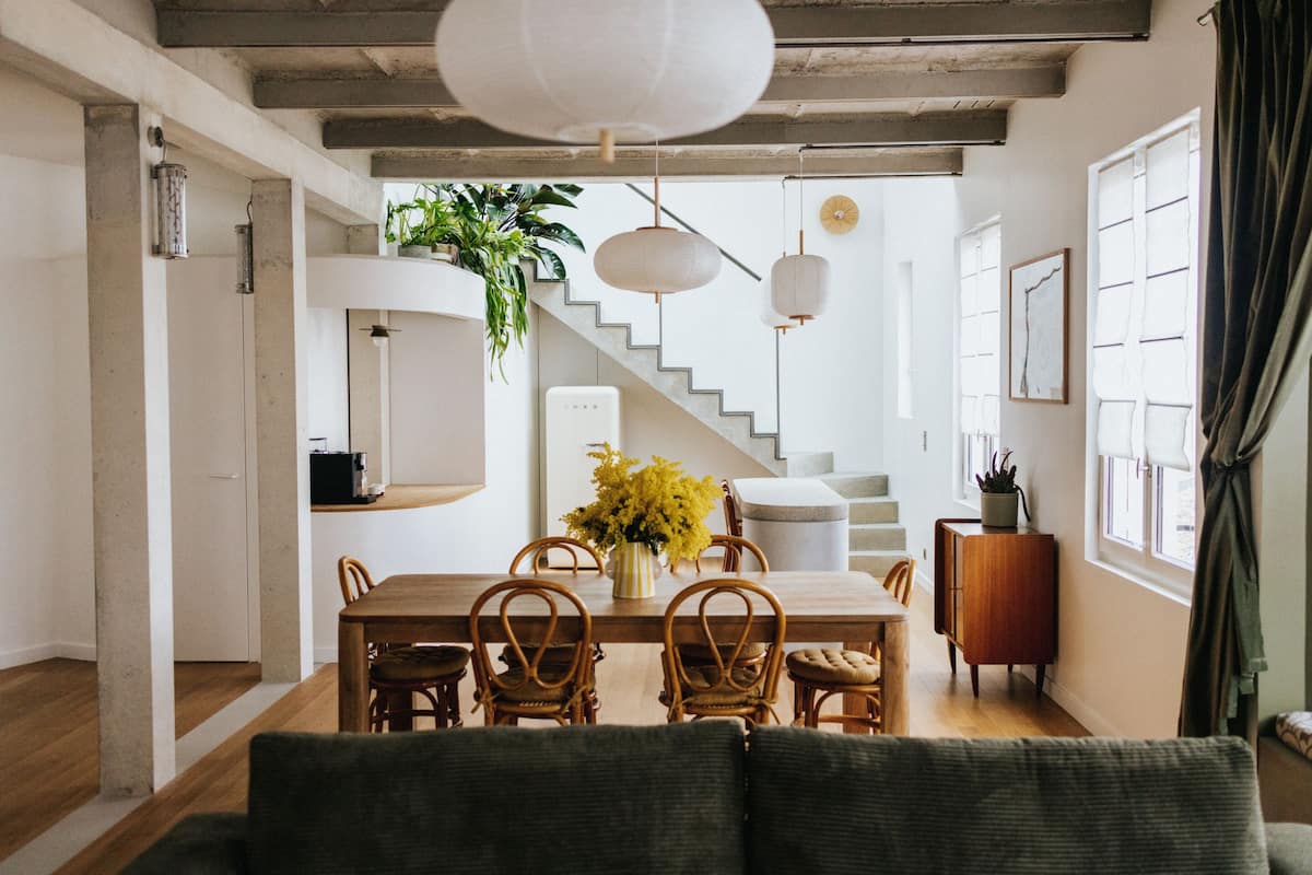 A dining area with a wooden table, wicker chairs, and a vase of yellow flowers. There is a staircase leading to another level, and a potted plant on the wall.