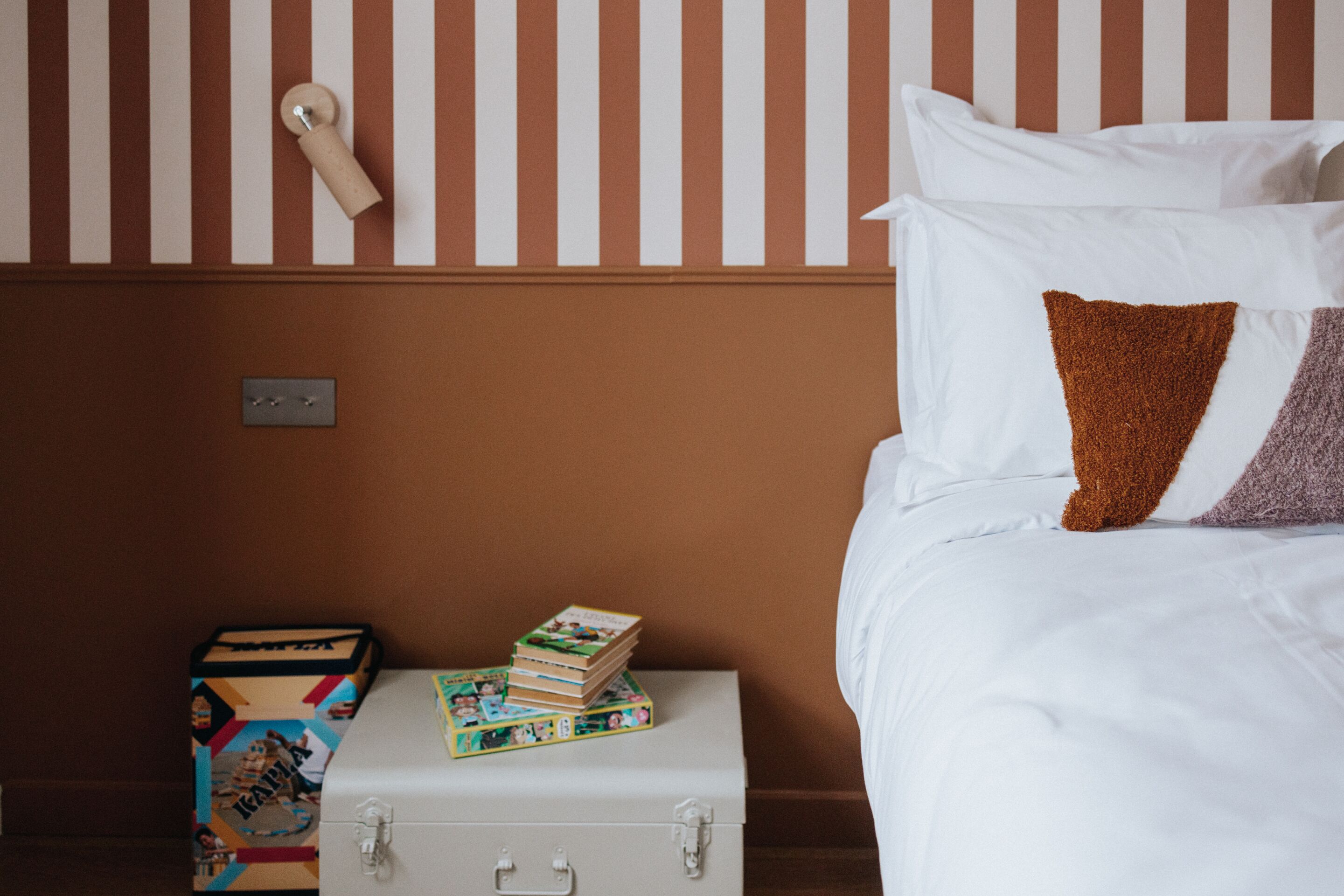 Una cama bien hecha con almohadas blancas y marrones, un cabecero a rayas y una maleta con libros a un lado.