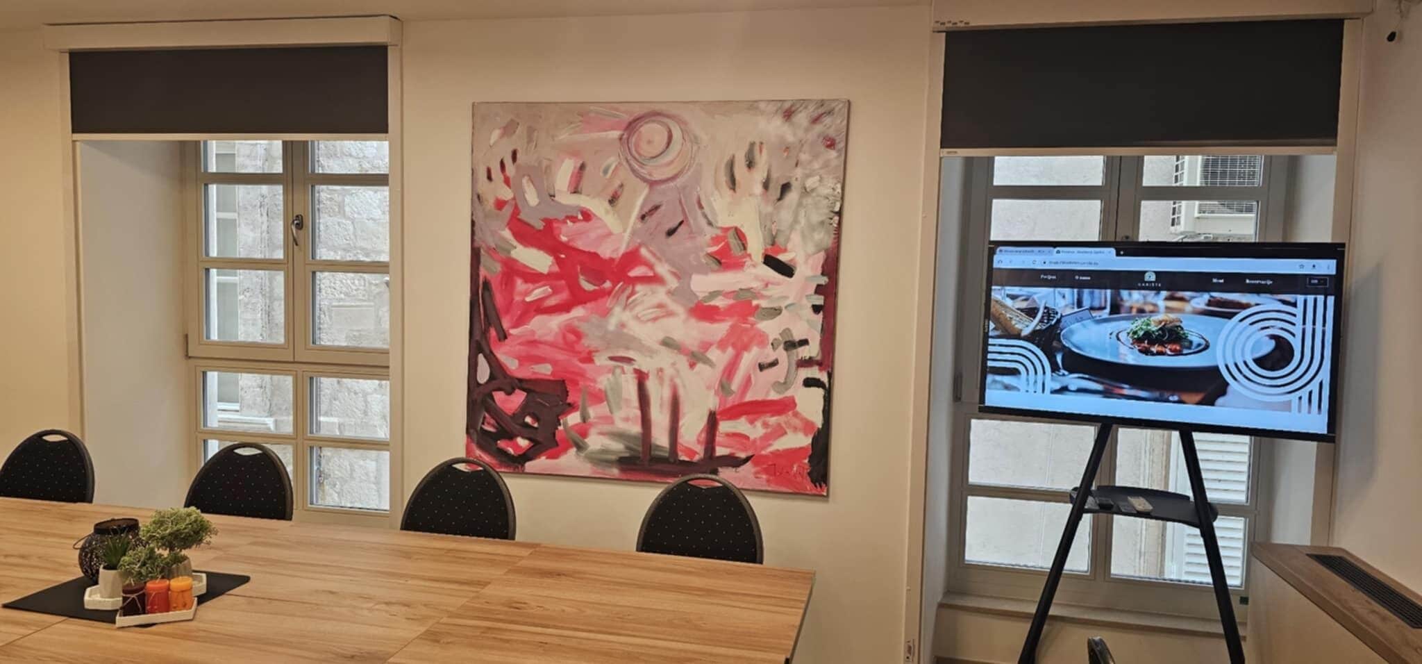 A conference room with a wooden table, chairs, a large abstract painting, and a projection screen displaying a restaurant scene.