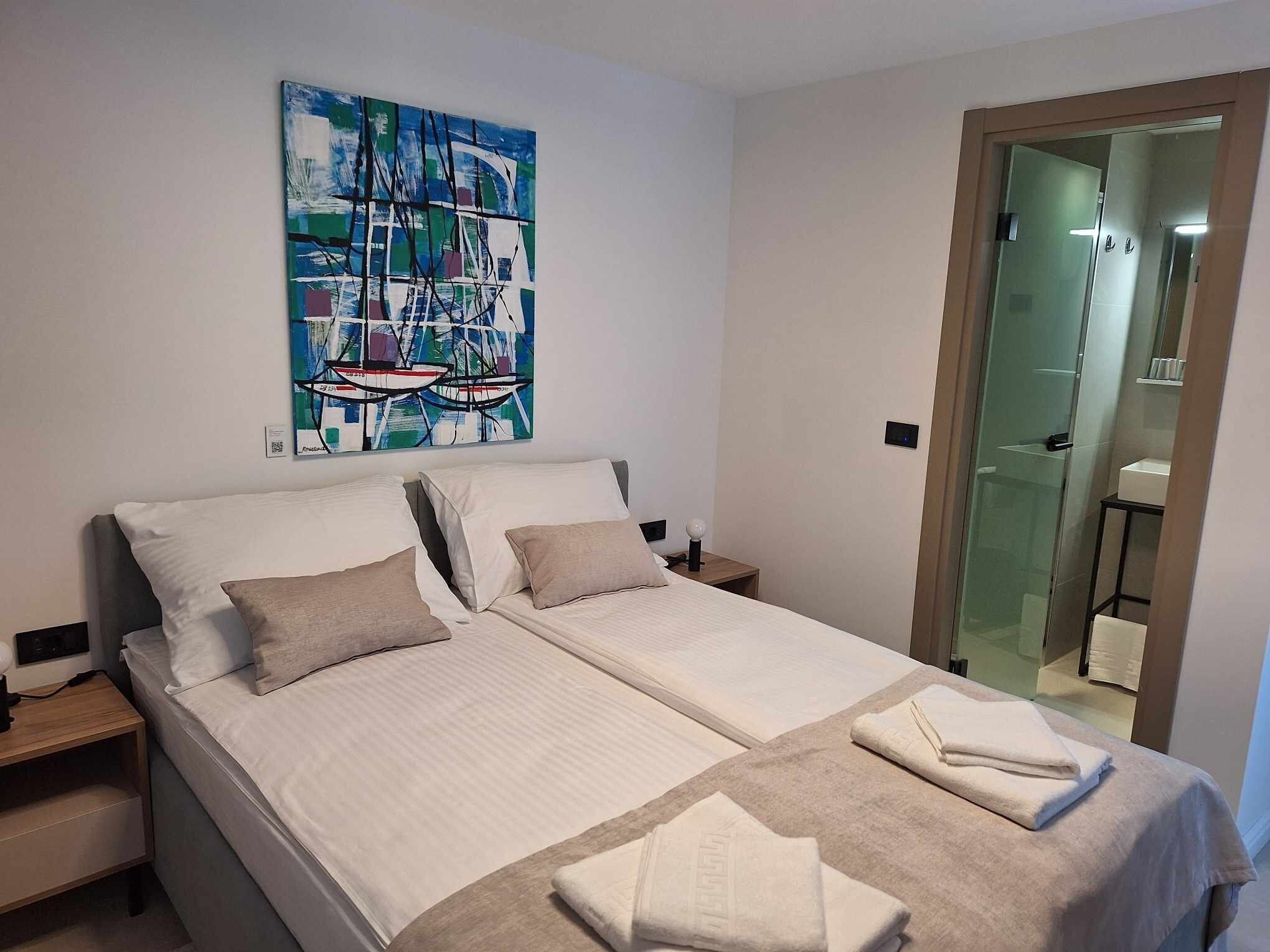 A double bed with two pillows, a bedside table, a nightstand, a painting of boats, and a bathroom visible through a glass door.