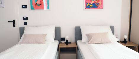 Habitación de hotel con dos camas, cada una con ropa blanca y una almohada rosa claro. Hay dos mesitas de noche con lámparas, y las paredes están decoradas con obras de arte coloridas.