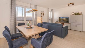 Una sala de estar moderna con una mesa de comedor de madera, sillas tapizadas en azul, un sofá gris y un televisor sobre un mueble.