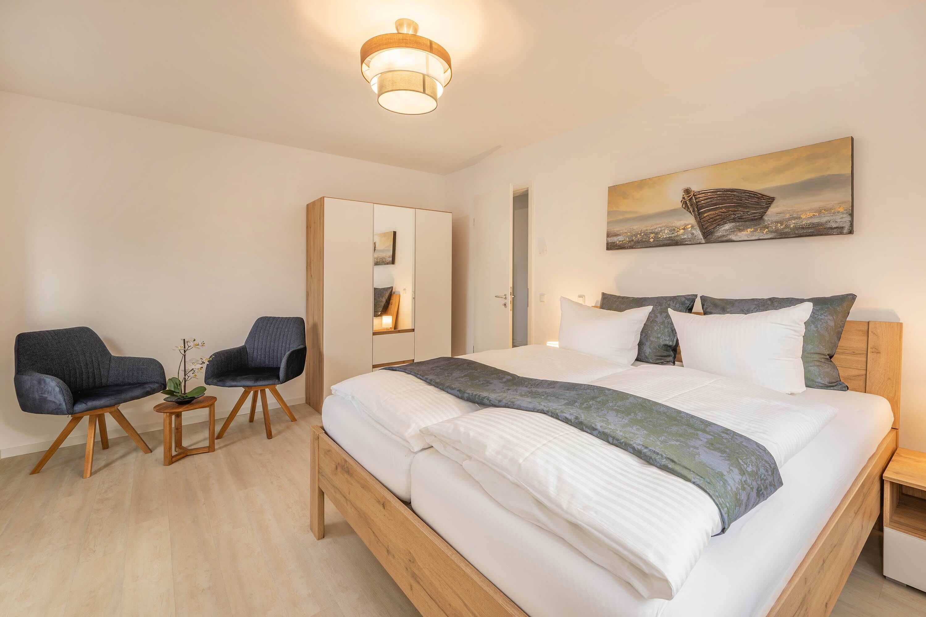 Een slaapkamer met een bed, twee stoelen, een kleintje tafeltje met een plant, een kledingkast en een schilderij aan de muur.