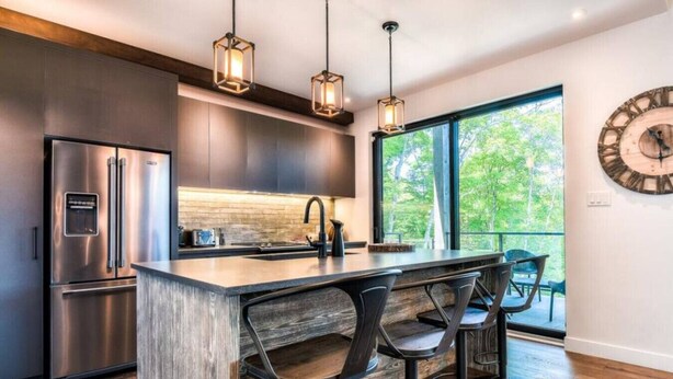 A modern kitchen with a stainless steel refrigerator, a wooden island with bar stools, pendant lighting, and a large window with a view of trees.