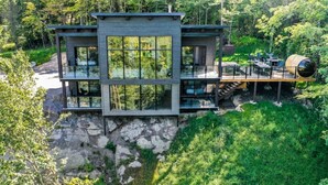 A modern multi-story house with large windows, situated on a rocky cliff surrounded by dense forest.