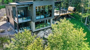A modern multi-story house with large glass windows, a deck, and a forested backdrop.