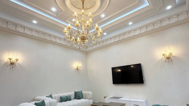 A spacious living room with a white sectional sofa, a teal ottoman, a gold-framed coffee table, and a large flat-screen TV mounted on the wall.