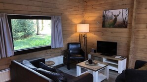 Een gezellig interieur van een chalet, met een leren bank, een zwarte fauteuil, een televisie en een schilderij van een hert op de muur.