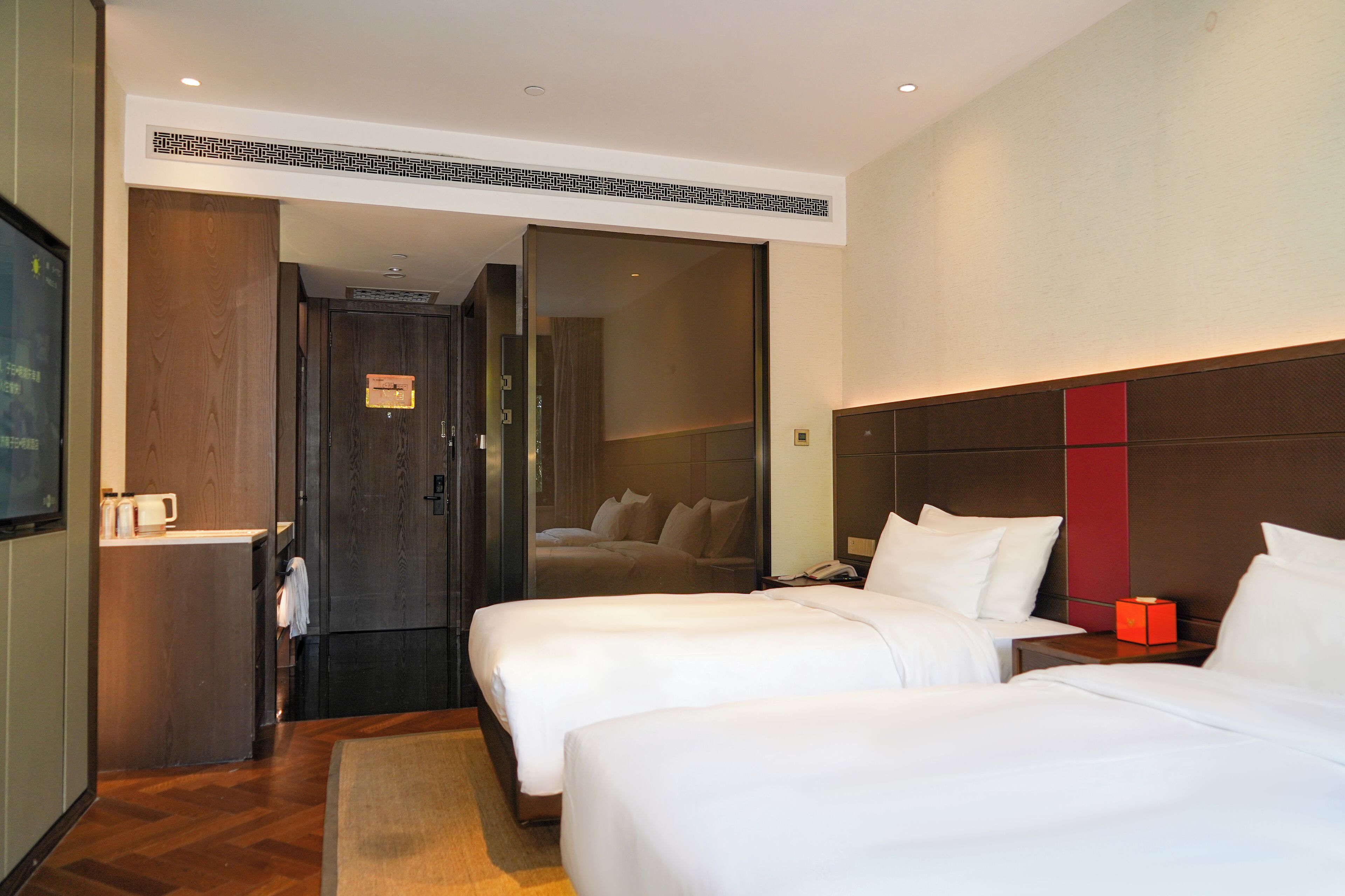 A hotel room with two beds, a wooden door, a small desk, and a nightstand with a red lamp.