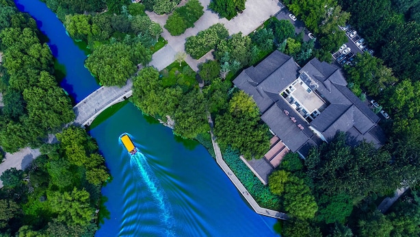 Vue aérienne d’une propriété riveraine dotée d’un quai pour bateaux, entourée d’une végétation luxuriante et bordée d’un immeuble résidentiel à toit en pente.