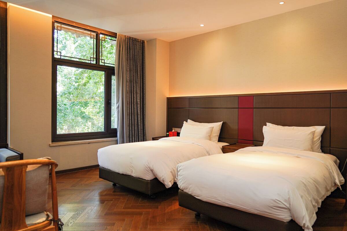 Een hotelkamer met twee bedden, een houten vloer, een raam met gordijnen en een nachtkastje met een lamp.