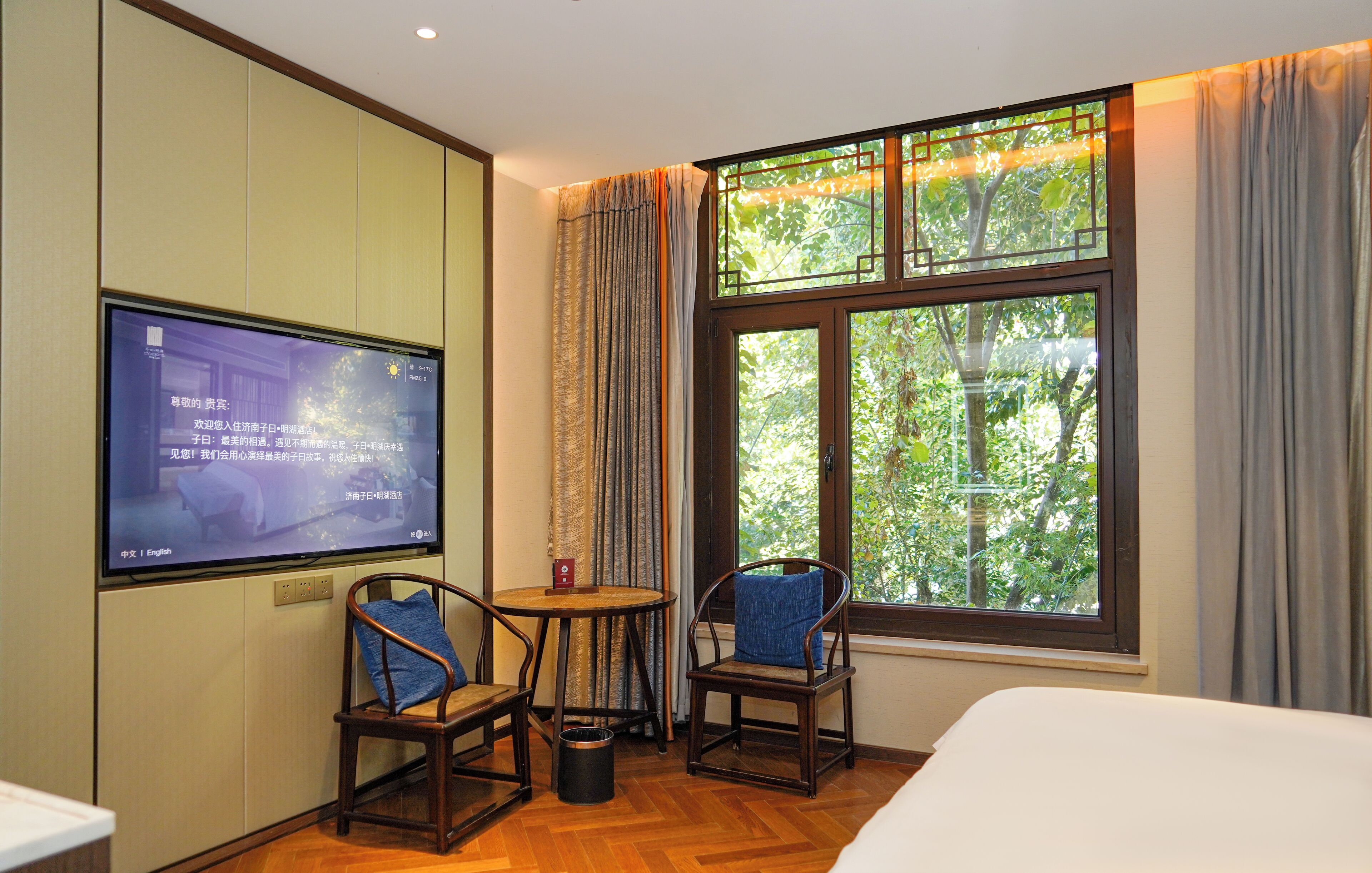 Une chambre d’hôtel avec un lit, deux chaises en bois, une petite table, un téléviseur fixé au mur et une grande fenêtre avec des rideaux.