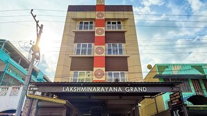 Entrada do hotel Lakshminarayana Grand, com uma placa, uma escadaria que leva à entrada e céu azul acima.