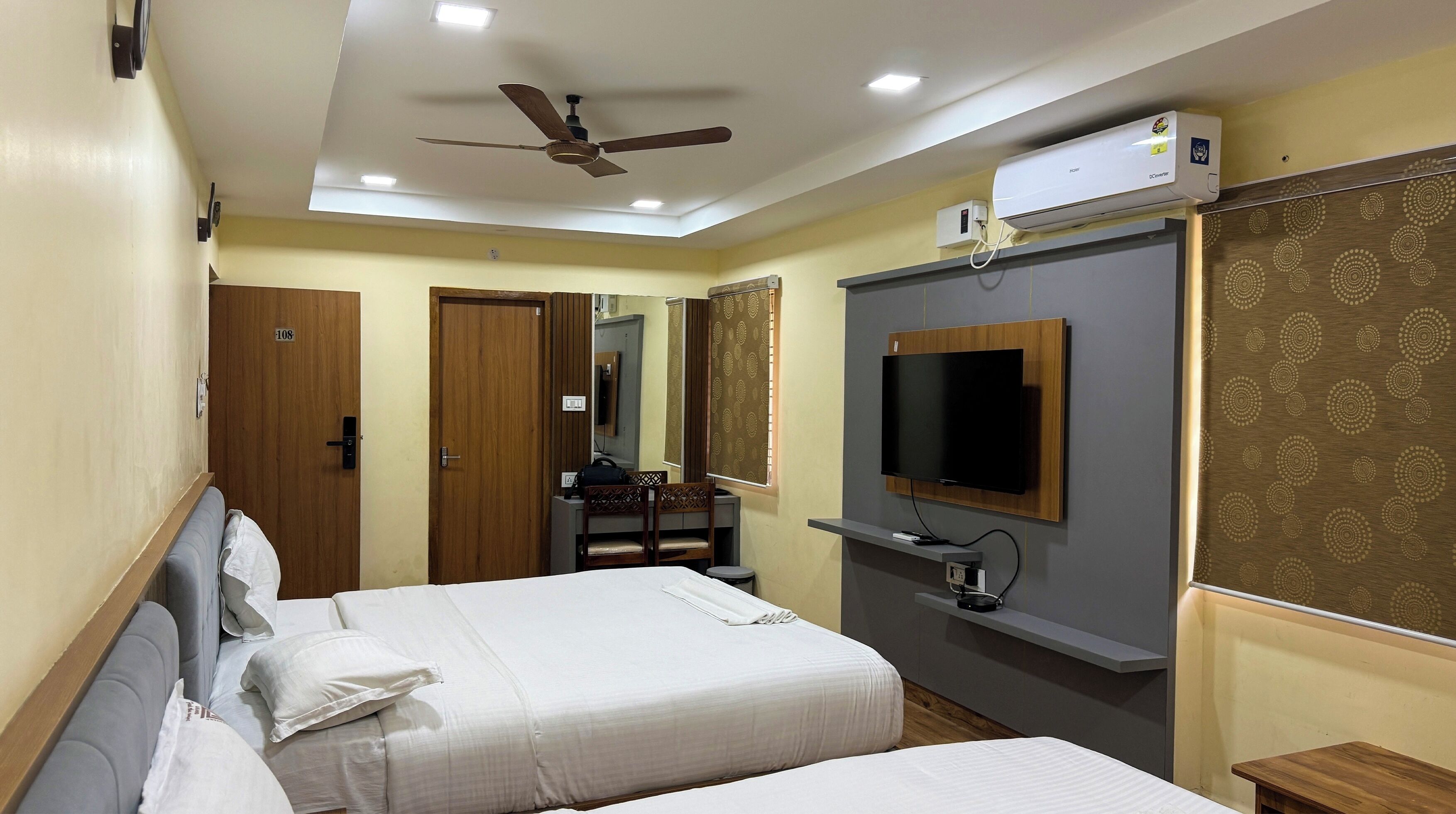 A hotel room with a bed, a desk with a chair, a TV mounted on the wall, a ceiling fan, and an air conditioning unit.
