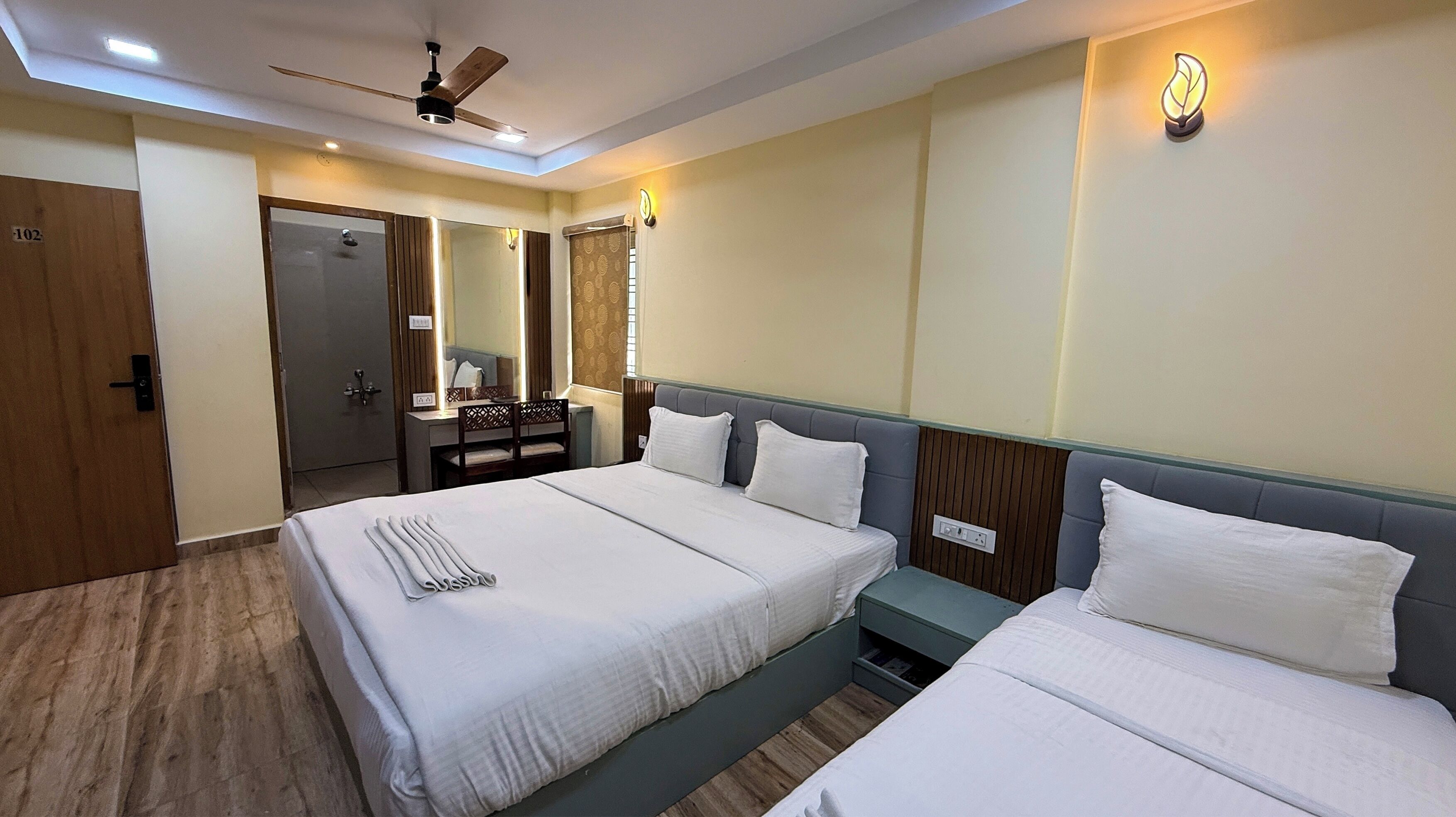 A hotel room with a large bed, a smaller bed, a desk with a chair, a mirror, and a ceiling fan.