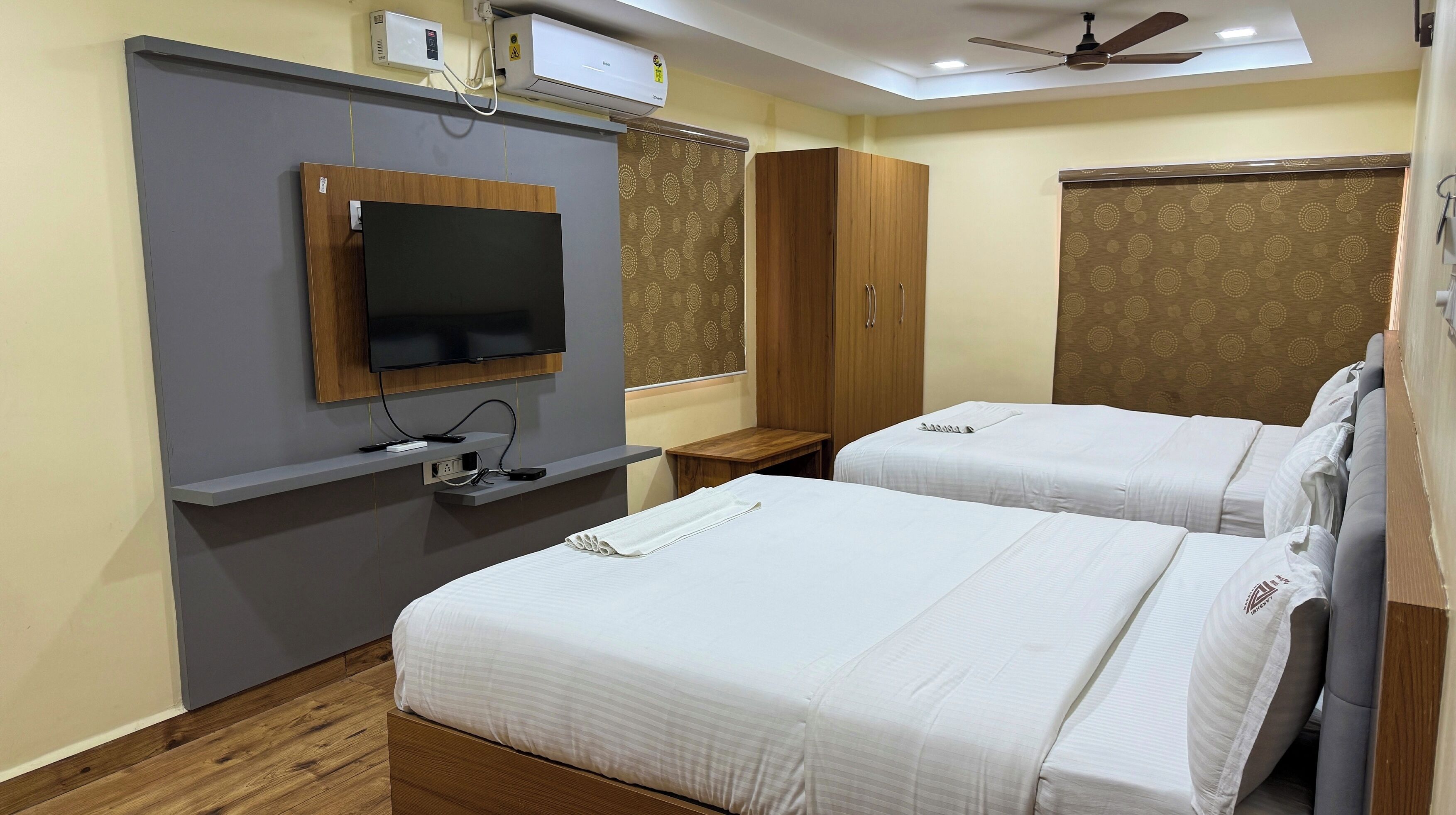 A hotel room with two beds, a TV mounted on the wall, a ceiling fan, and a window with patterned curtains.