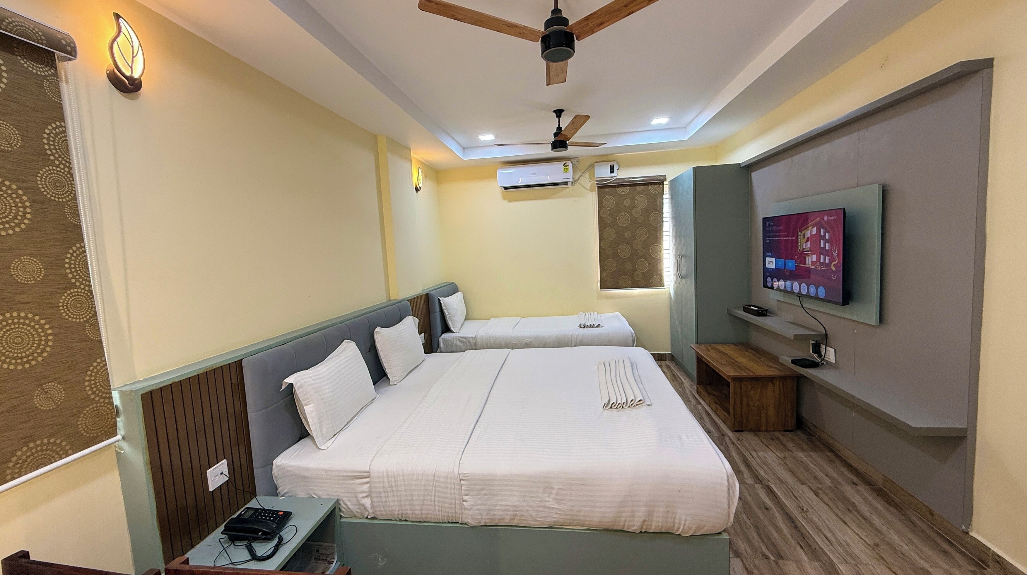 A hotel room with two beds, a ceiling fan, an air conditioning unit, a television, and a small wooden table.