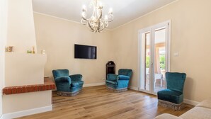 Ein geräumiges Wohnzimmer mit Holzboden, zwei smaragdgrünen Sessel, ein beiges Sofa, einem Flachbildfernseher und einer Kandelaber.