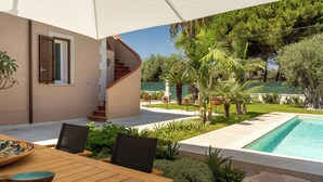 Ein Holztisch mit Stühlen und einer Schüssel, ein Schwimmbecken und eine Treppe, die zu einem überdachten Bereich mit Blick auf Bäume und einen grünen Zaun führen.