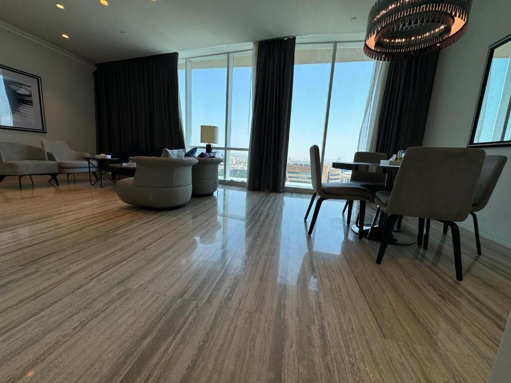 A spacious hotel room with a large window offering a city view, a modern chandelier, and a clean, polished wooden floor.