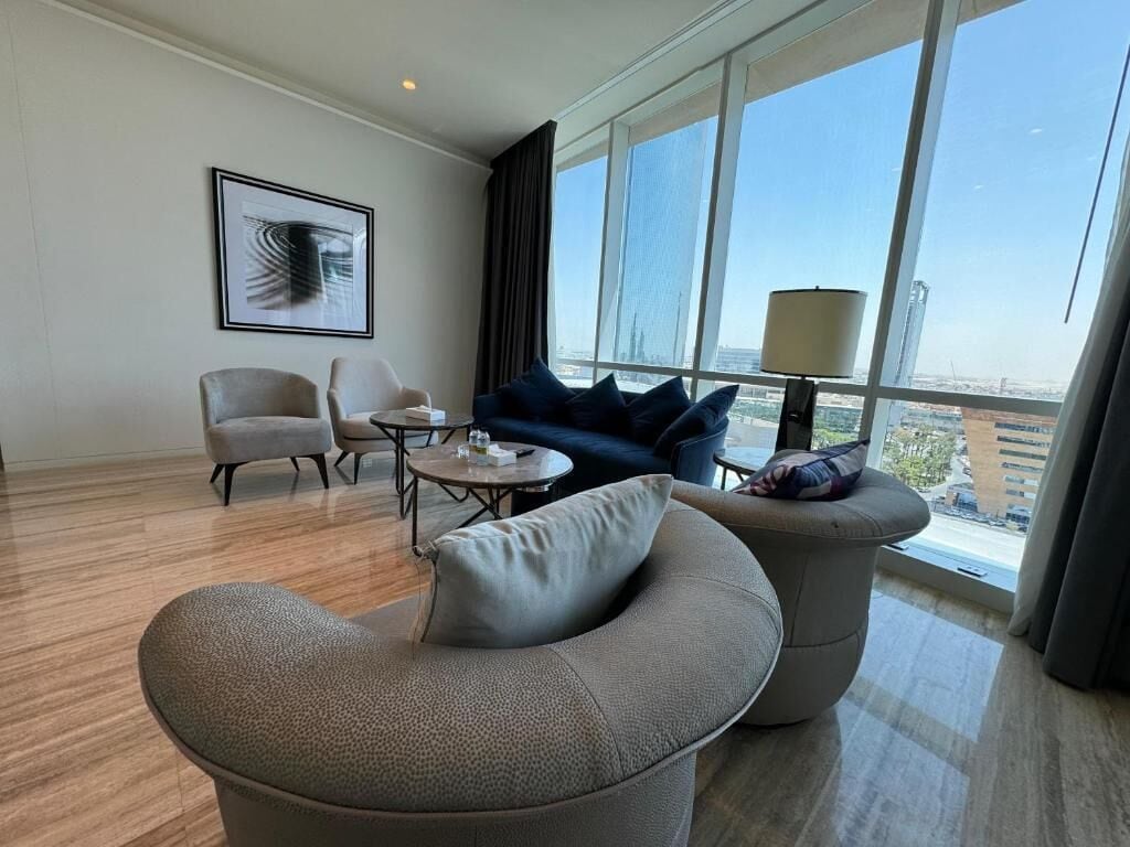 A modern hotel room with a large window offering a city view, a sofa, two armchairs, a coffee table, and a framed artwork on the wall.