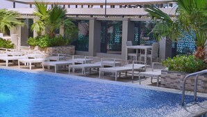 A swimming pool with lounge chairs and a pergola, surrounded by greenery and modern buildings. - Luxury Apartment in DAMAC Towers (Riyadh)
