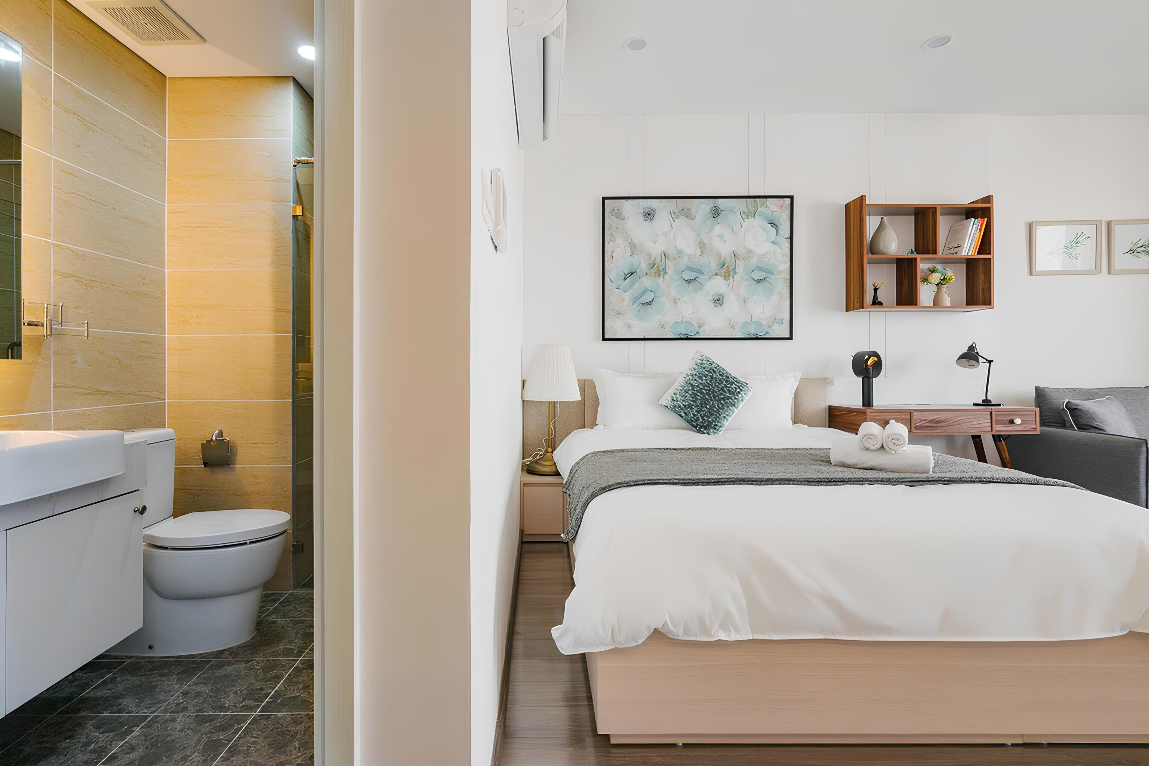 A modern hotel room with a bathroom, a bed with white linens, a bedside table with a lamp, and a bookshelf.