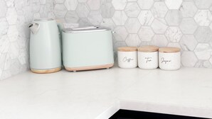 Une cuisine avec un pare-feu en marbre blanc, une bouilloire, un grille-pain et trois pots à épices étiquetés « café », « thé » et « sucre ».