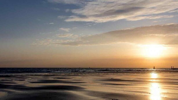 Una spiaggia al tramonto con mare calmo e un faro in lontananza.